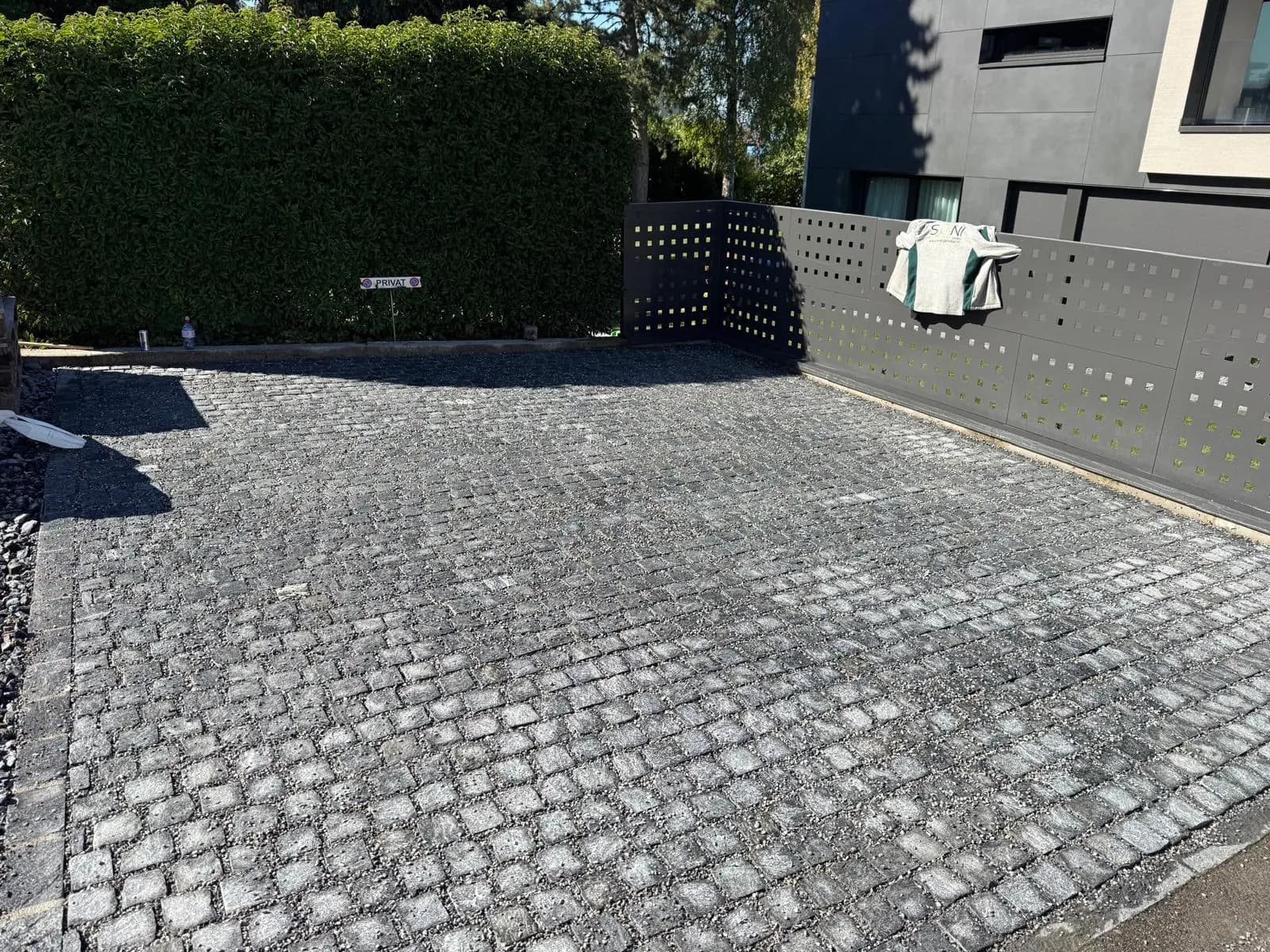 Fertig verlegte Einfahrt aus Granit-Kleinpflaster mit offenen Sickerfugen, eingefasst von Sichtschutzzaun und Buchenhecke – Soni Gartenbau