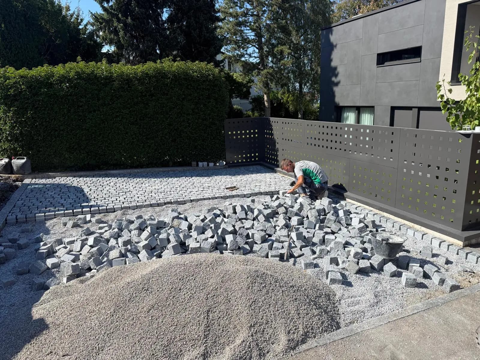 Baustelle eines Vorplatzes: Mitarbeiter verlegt Granitpflaster im Splittbett, daneben der Steinvorrat, im Hintergrund ein modernes Wohnhaus – Soni Gartenbau
